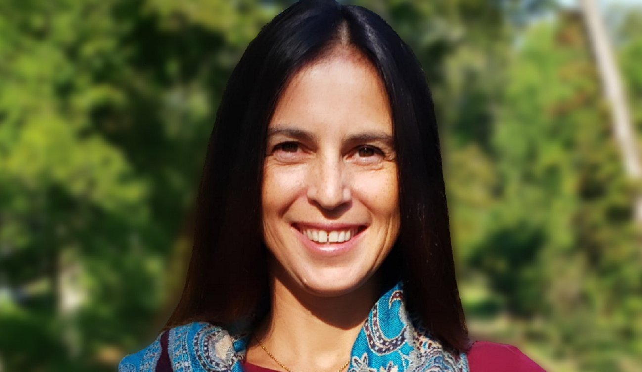 Jessica Bulling smiles for the camera, wearing a red shirt and a blue scarf. It is a sunny day and there are green trees in the background. 